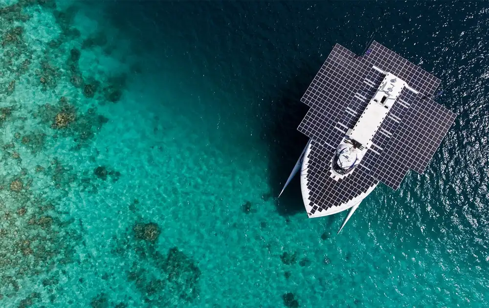 Le catamaran Porrima vu du ciel, son pont couvert de panneaux photovoltaïques, au mouillage au-dessus d'un récif corallien.